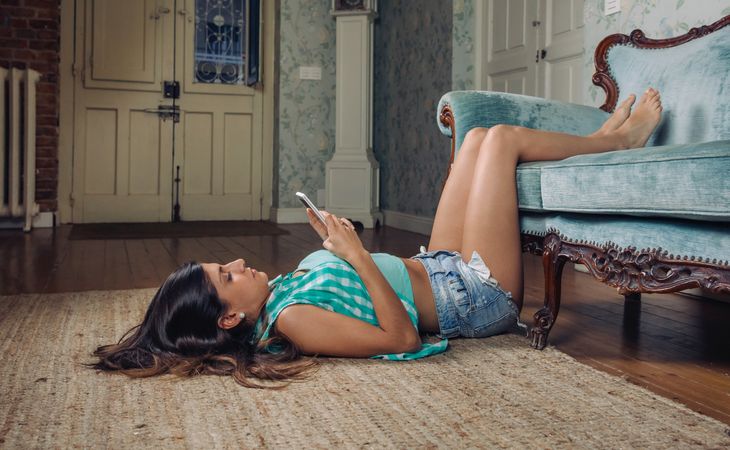 Woman lies on floor with feet resting on sofa checking phone