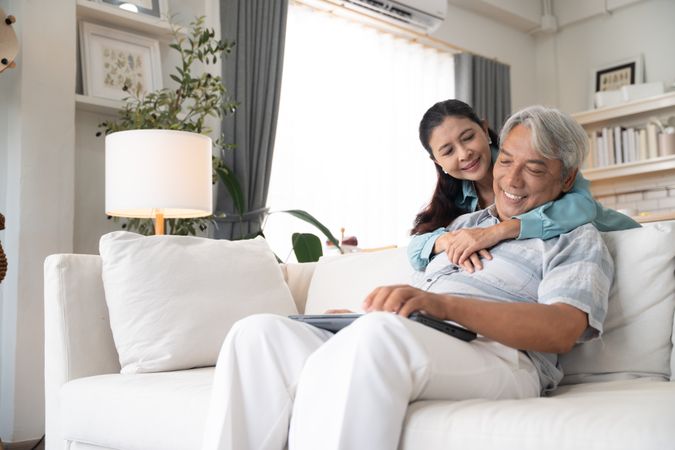 Asian man sitting on couch working on laptop while his wife embraces