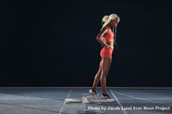 Female sprinter standing beside a starting block on running track on a dark background 5wmpyb