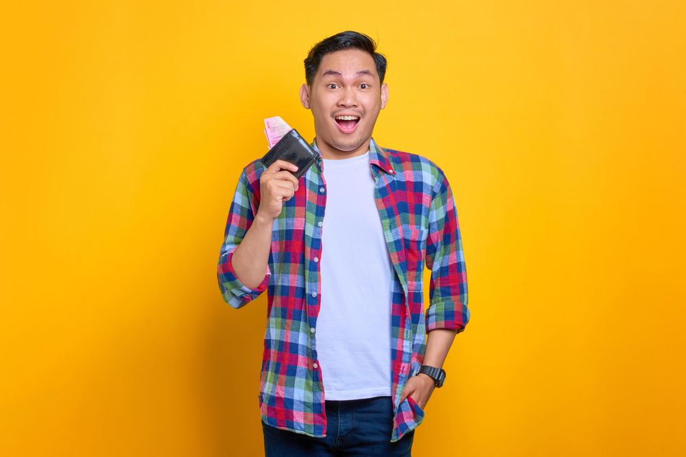 Excited Asian man smiling while holding wallet with hand in his pocket
