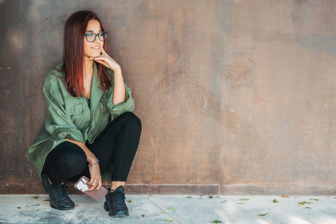 Stylish confident bright woman in glasses leaning on wall and looking at camera