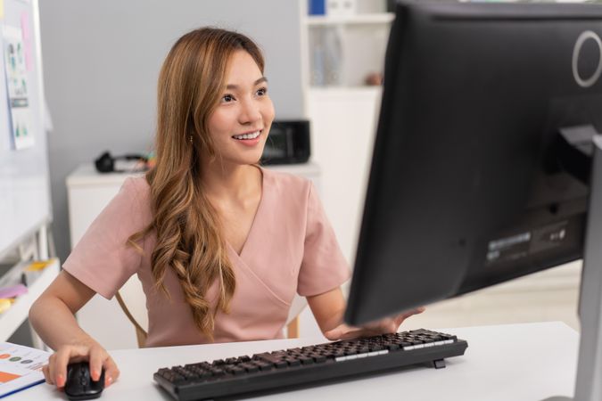Confident Asian woman sitting in office and using computer while looking at monitor