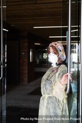 Smiling nurse in PPE holds glass door open while looking at camera beXDK4