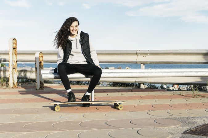 Teenage girl sitting on the bench smiling with her feet on the longboard in front of the sea