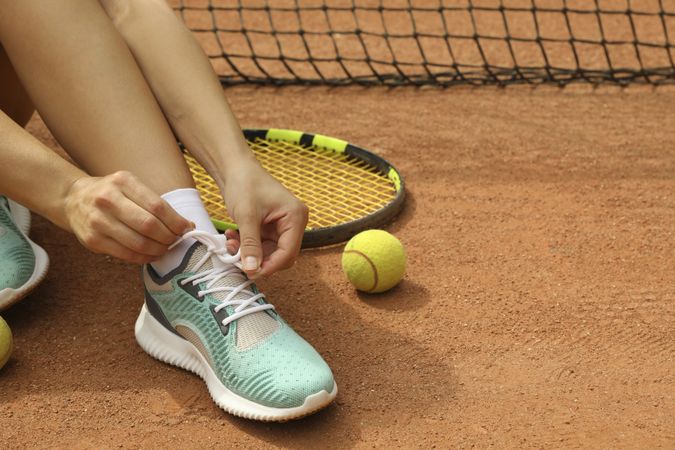 Woman tying shoelaces on clay court with racket and tennis ball