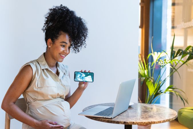Smiling Black pregnant woman showing baby ultrasound picture to laptop