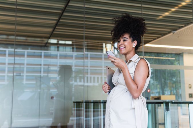 Smiling Black pregnant woman using smartphone and wireless earphones