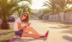 Young girl with skateboard and smartphone sitting outdoors on summer