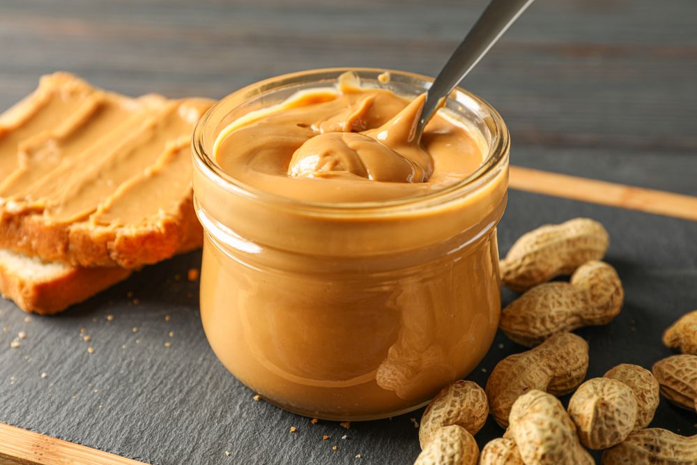 Glass jar with peanut butter and spoon, peanut, peanut butter sandwich and cutting board on wooden background, space for text and closeup