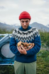 Man with tent wearing blue wool sweater cradling pile of foraged  mushrooms