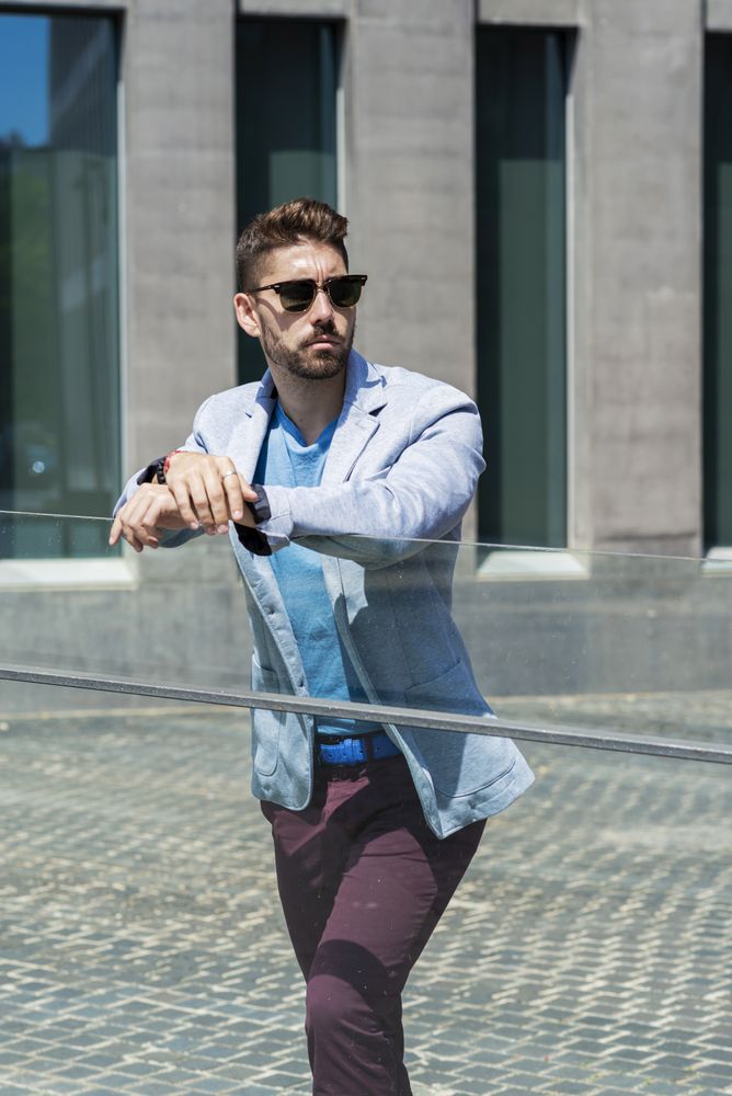 Portrait of a young bearded man with sunglasses leaning on glass railings outdoors in the city while looking aside