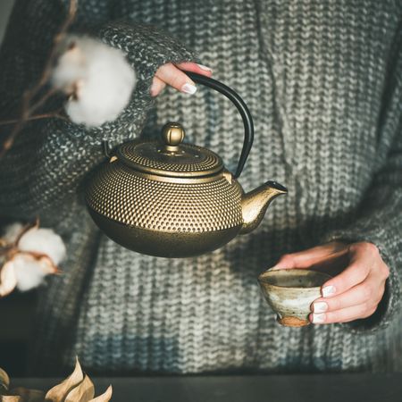 Woman in cozy sweater pouring from traditional Japanese tea set, with cotton, square crop