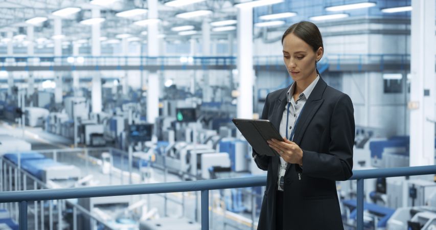 Thoughtful Female Engineer Monitoring and Analyzing Conditions at a Modern Tech Electronics Factory with Automated Robot Arms Working with the Help of Tablet Computer