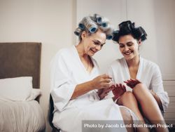 Two women in bathrobe with hair rollers sitting together at home doing their nails 4OpYgb