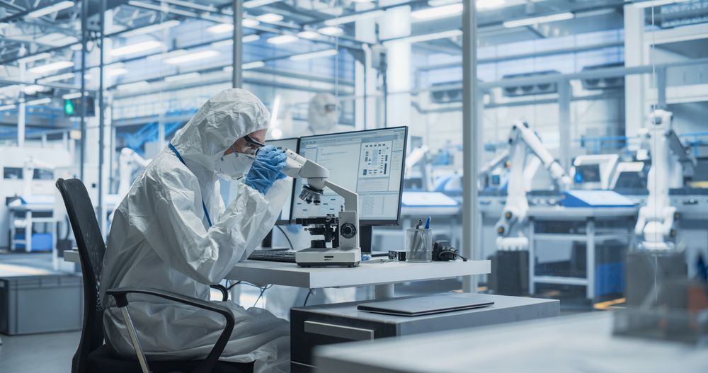 Modern Factory: Industrial Engineer Working on Desktop Computer in Clean Sterile Coveralls. Scientist Using a Microscope, Developing Advanced Solutions for High-Tech Medical Vaccine and Gene Research