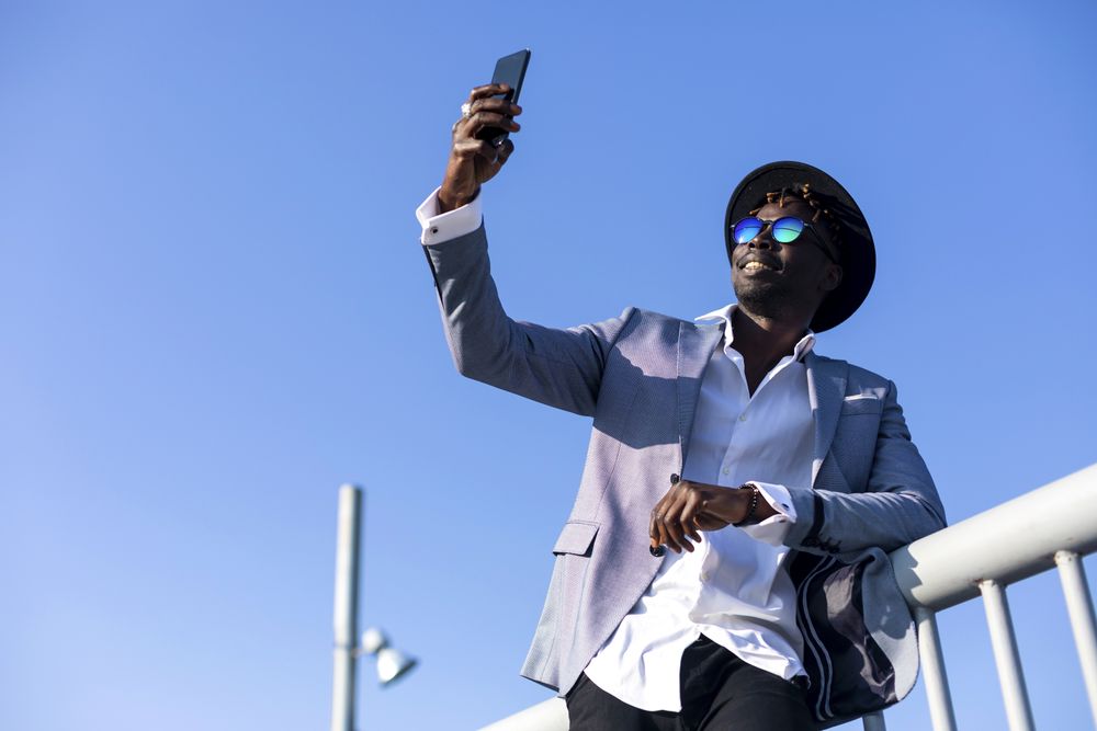 Black man in suit leaning on a bar taking a selfie on a sunny day