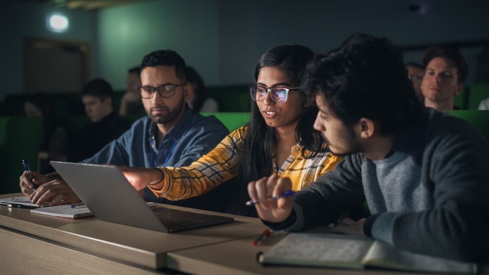 Group of Multiethnic Enthusiastic Students Collaborate in University Classroom, Compare Research Project Findings on a Laptop Computer.