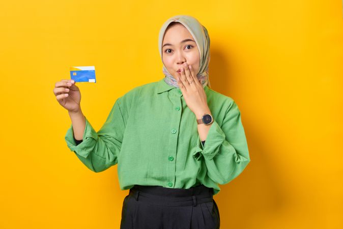 Muslim woman in headscarf and green blouse holding credit card and making surprised face