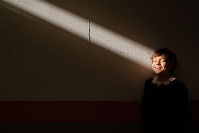 Smiling boy standing with closed eyes at wall in sunbeam