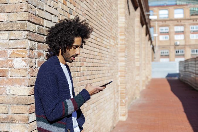 Side view of Black man looking down at his smartphone while leaning on a brick wall outdoors on sunny day