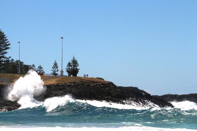 Waves crash against rocky shore with people observing from above on a sunny day in coastal area