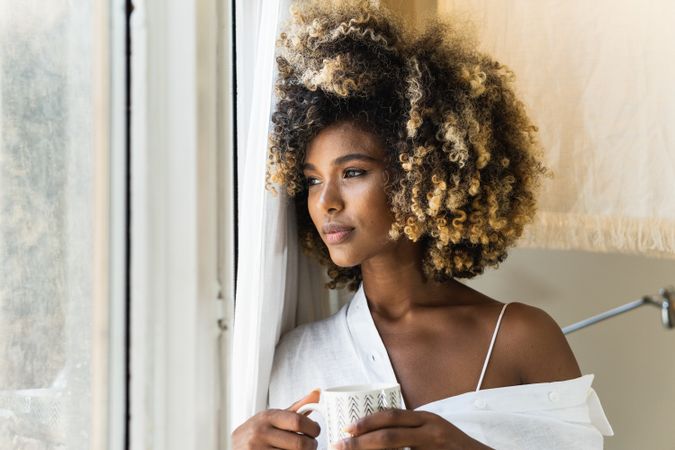 Dreamy Black woman with cup of drink near window