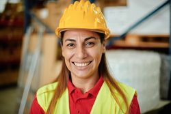 Portrait of a happy adult working woman looking at the camera inside a warehouse wearing a hard hat and safety clothing - Focus on the Face