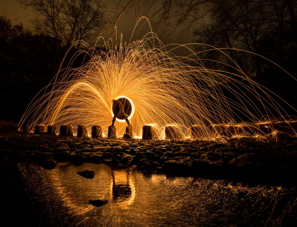 Person with burning circle on stones between water near hills at night