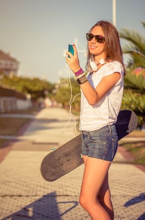 Young girl with skateboard and headphones looking smartphone