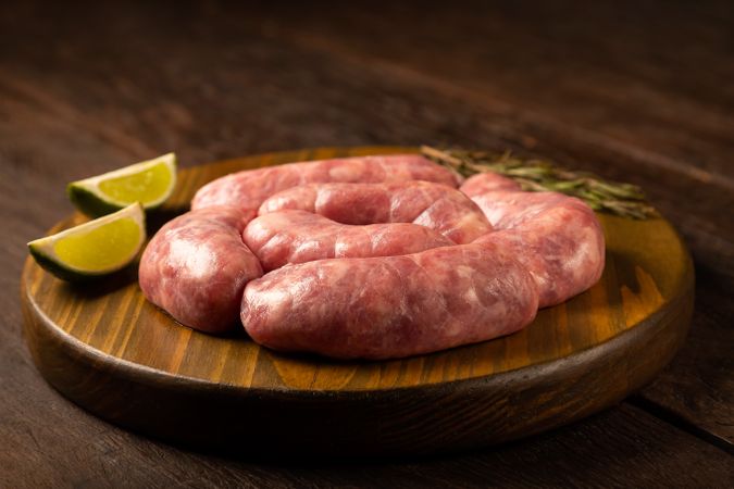 Raw sausages arranged on wooden board with rosemary and lime