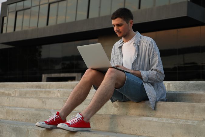 Young man sits in steps and working in laptop outdoor. Freelancer