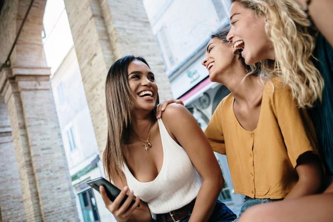 Laughing female friends sitting in Italian city and laughing at something on phone