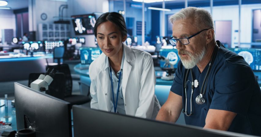 Asian Female Scientist Using Desktop Computer And Talking To White Male Doctor In Medical Hospital Research Lab.