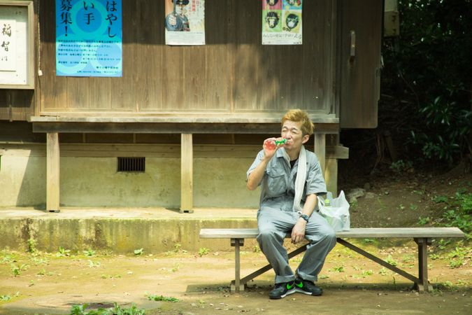 Man in work suit drinking juice and sitting beside plastic bag on wooden bench outdoor