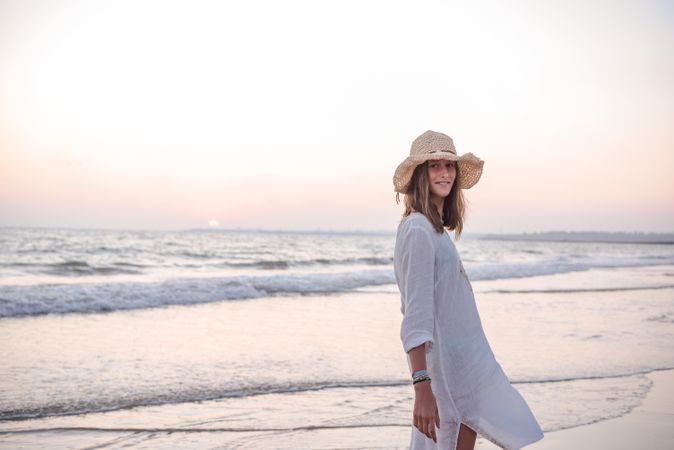 Charming woman in light white dress looking at camera over shoulder on wavy beach