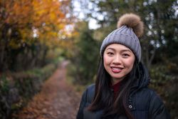 Smiling Asian woman in warm clothes standing in park with trees and stony fence