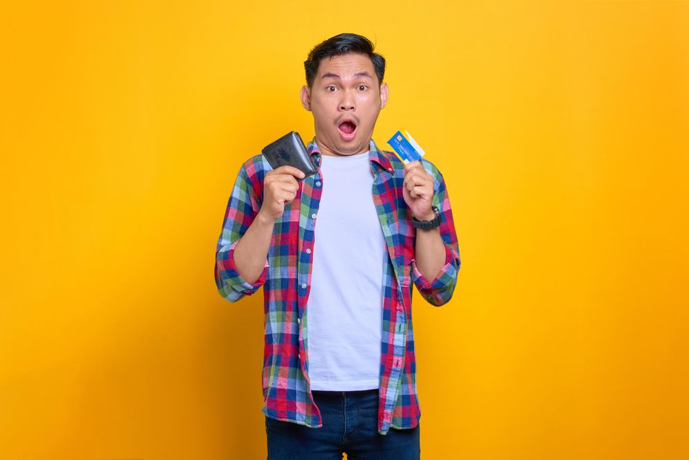 Surprised Asian man smiling while holding wallet with credit card in his other hand