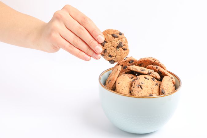 Young woman holding tasty chocolate chip cookie over bowl on blank background, closeup
