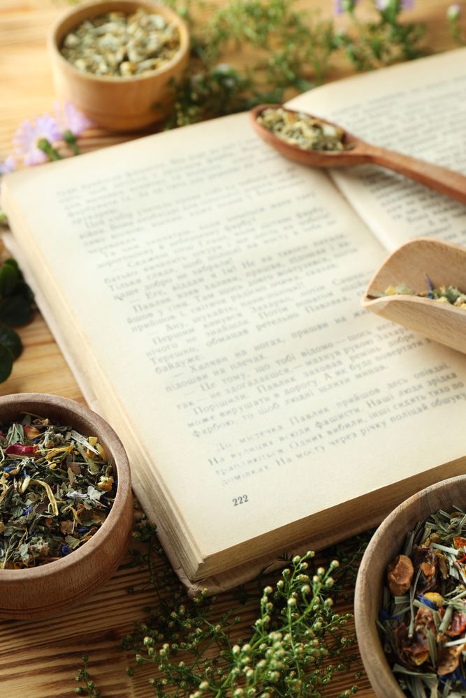 Book, herbs in bowls and scoops on wooden background, close up