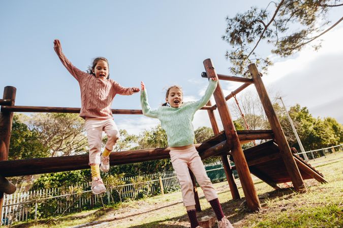 Happy little twin sisters jumping off a wooden log at the park