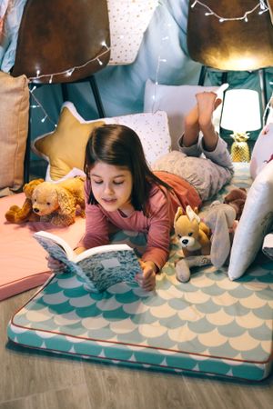 Portrait of serene little girl looking book while resting surrounded by cushions in homemade teepee
