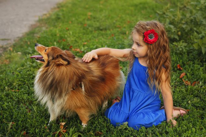 Happy child in blue dress with flower in hair petting dog in the grass