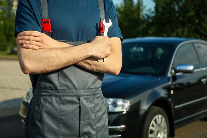 Man in overalls holding a wrench with folded arms. Car inspection