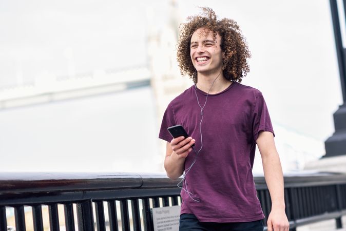 Curly haired man smiling and listening to music on smartphone along river walk