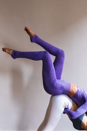 Anonymous flexible woman supporting female friend on back while doing Eka Pada Adho Mukha Svanasana yoga pose