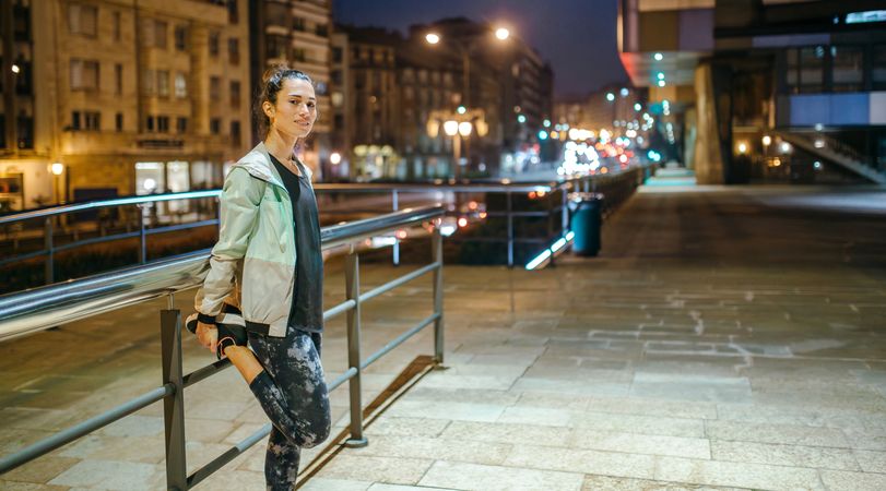 Fit woman stretching her legs next to banister before training at night in city