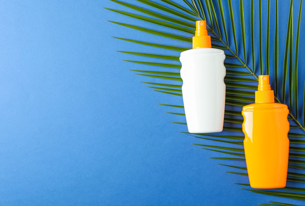 Composition with sunscreen sprays and palm leaf on color background, space for text