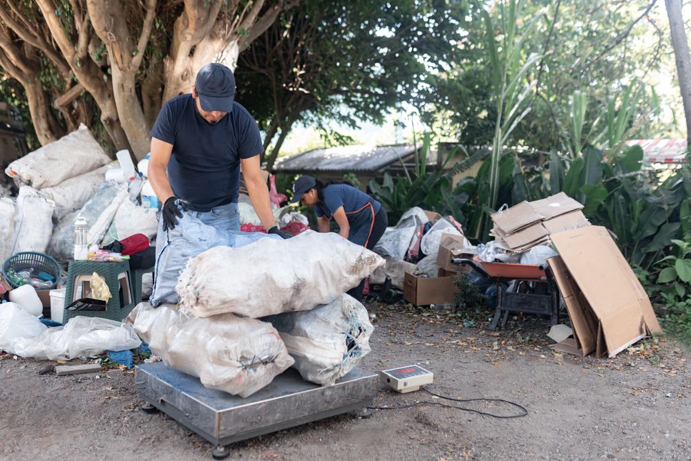 An adult Hispanic man weighs a pile of sacks of reclaimed materials, supported by a woman in the background