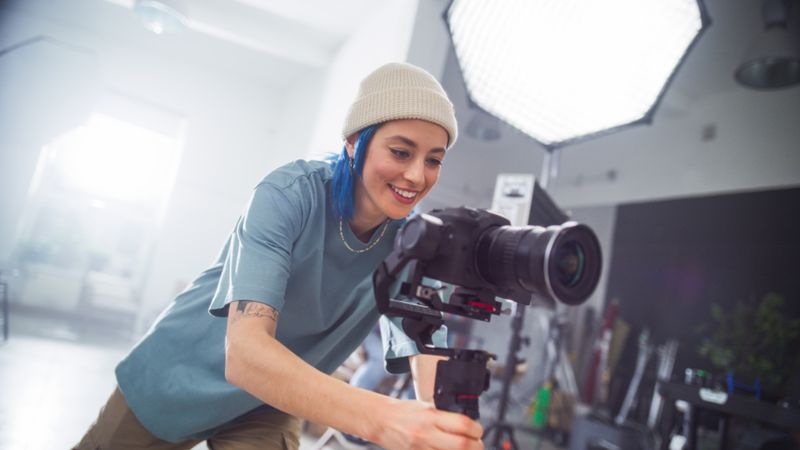 Focused Young Female Videographer Adjusts Professional Camera In Well-lit Studio, Capturing Essence Of Modern Filmmaking And Creativity In Advertising Production.