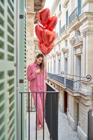Cheerful woman with bunch of red heart balloons on balcony